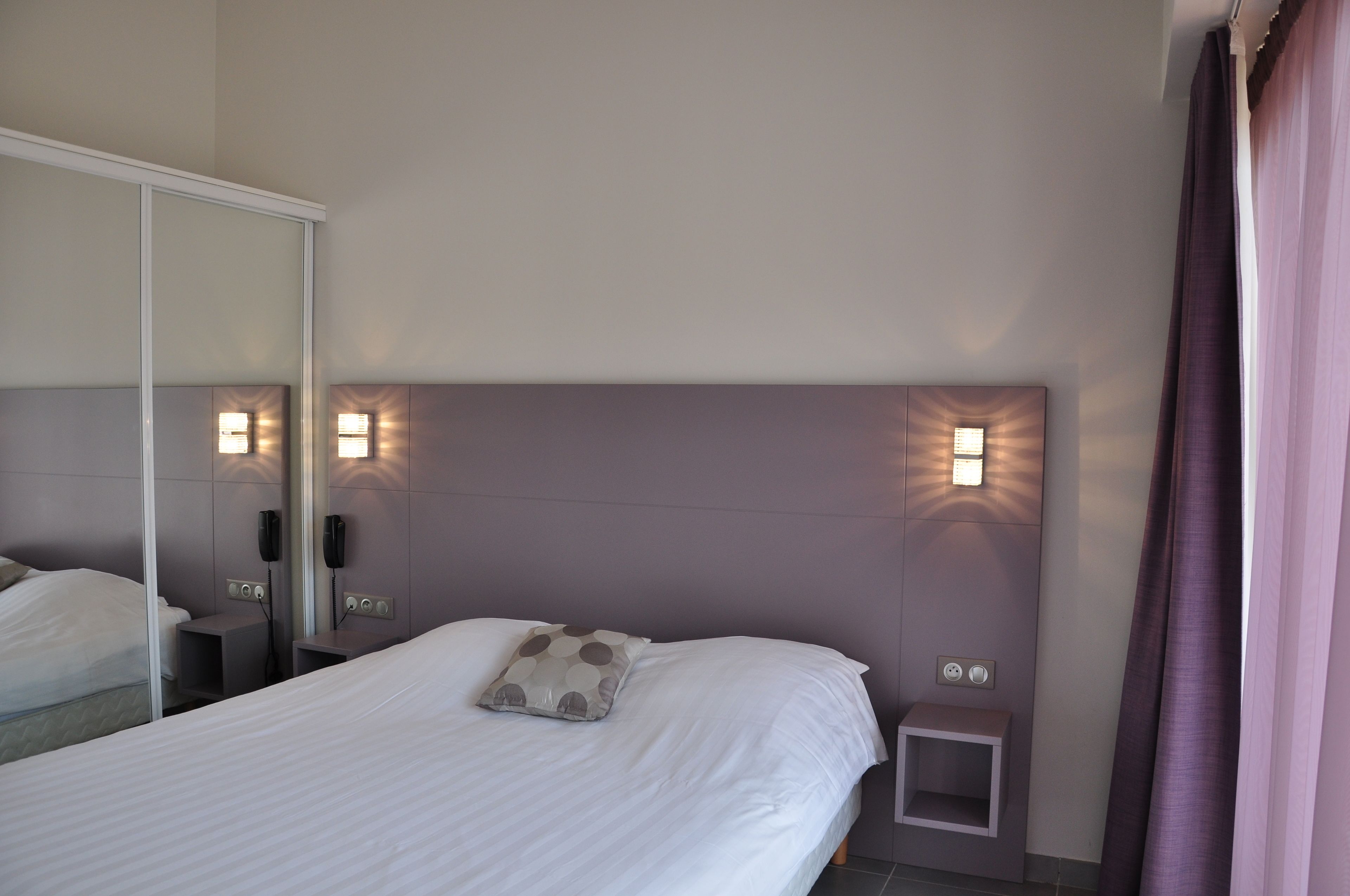 A hotel room with a large bed, bedside tables, a mirror, and wall-mounted lights.