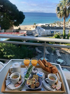 Ein Frühstücksbrett mit Gebäck, Saft und Danone-Joghurt auf einem Balkon mit Blick auf einen Strand.