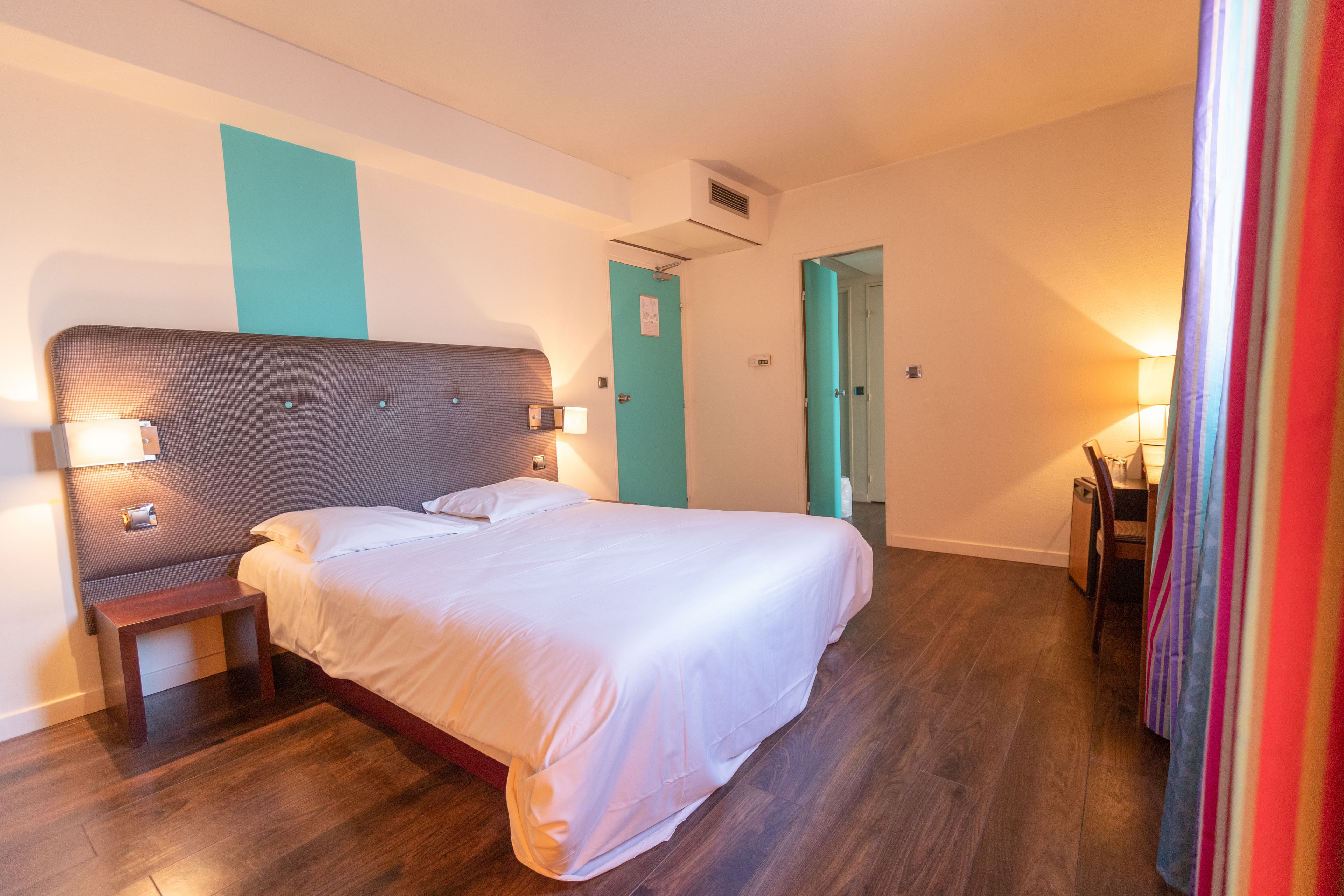 A hotel room with a large bed, a bedside table, a nightstand, a desk with a chair, and a door leading to another room.