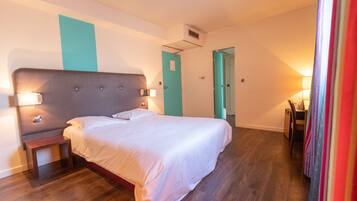 A hotel room with a large bed, a bedside table, a nightstand, a desk with a chair, and a door leading to another room.