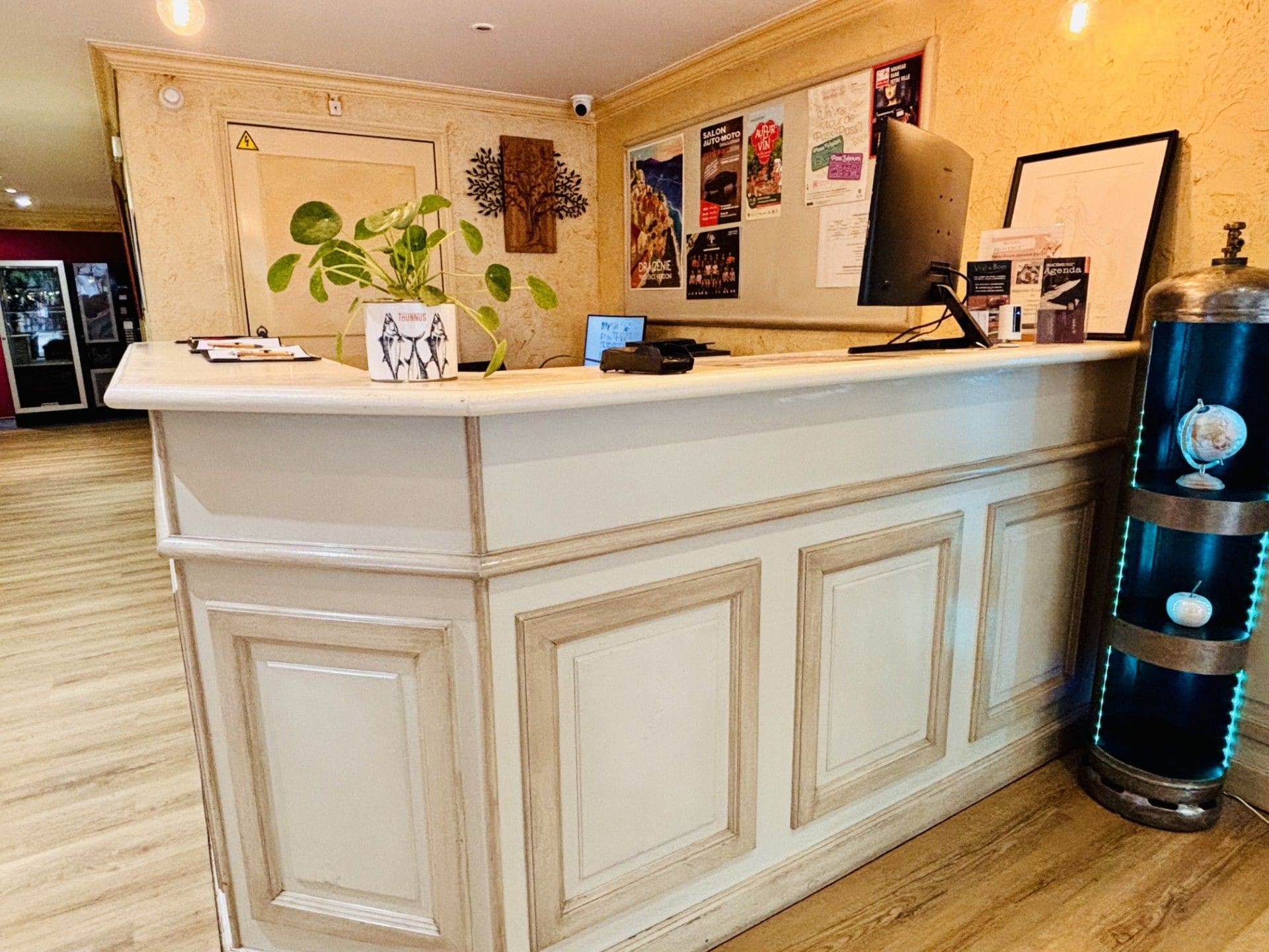 Un comptoir d’accueil avec une plante en pot, un ordinateur et divers affiches accrochées au mur.