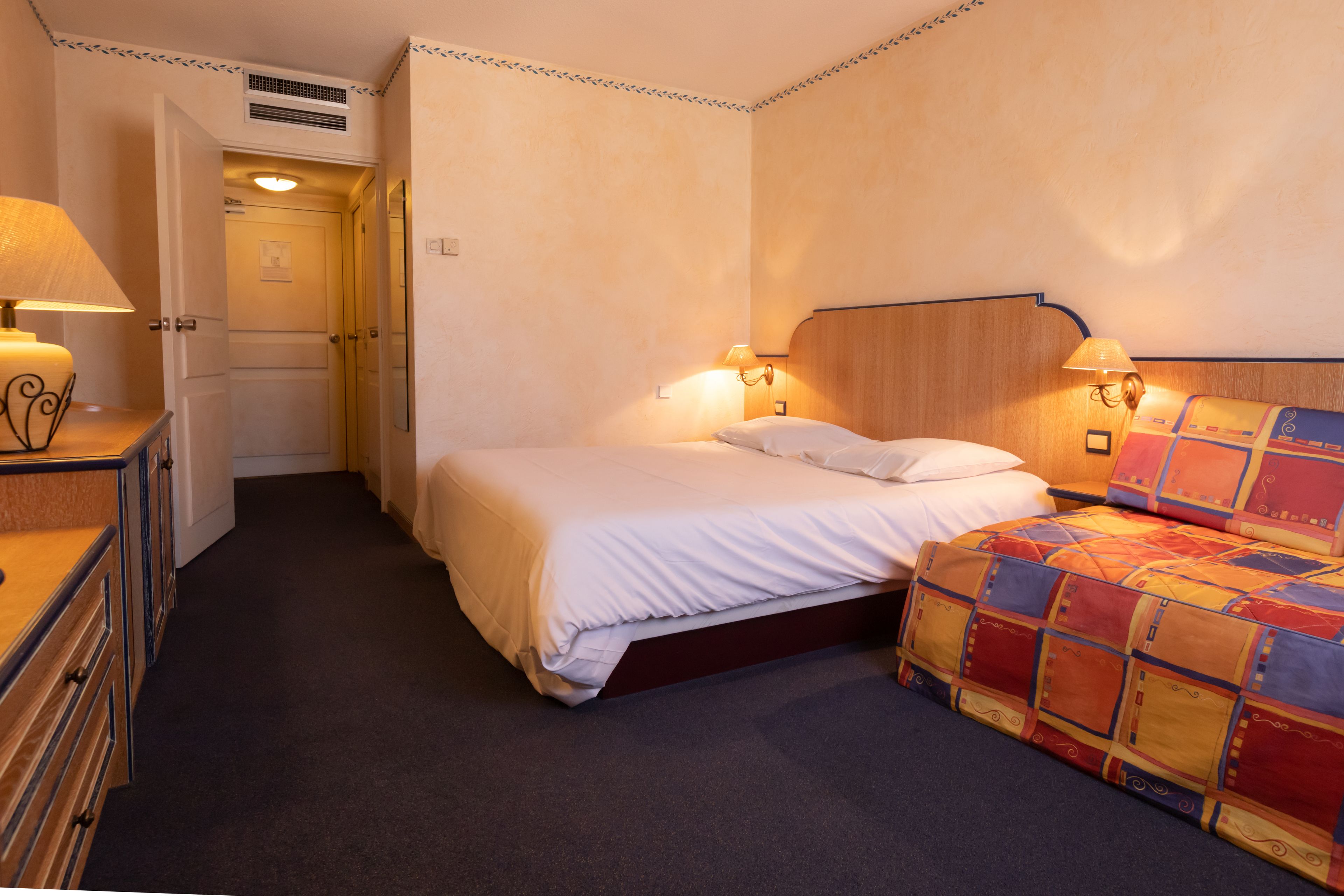 A hotel room with a bed, bedside lamps, a dresser, and a door leading to another room.