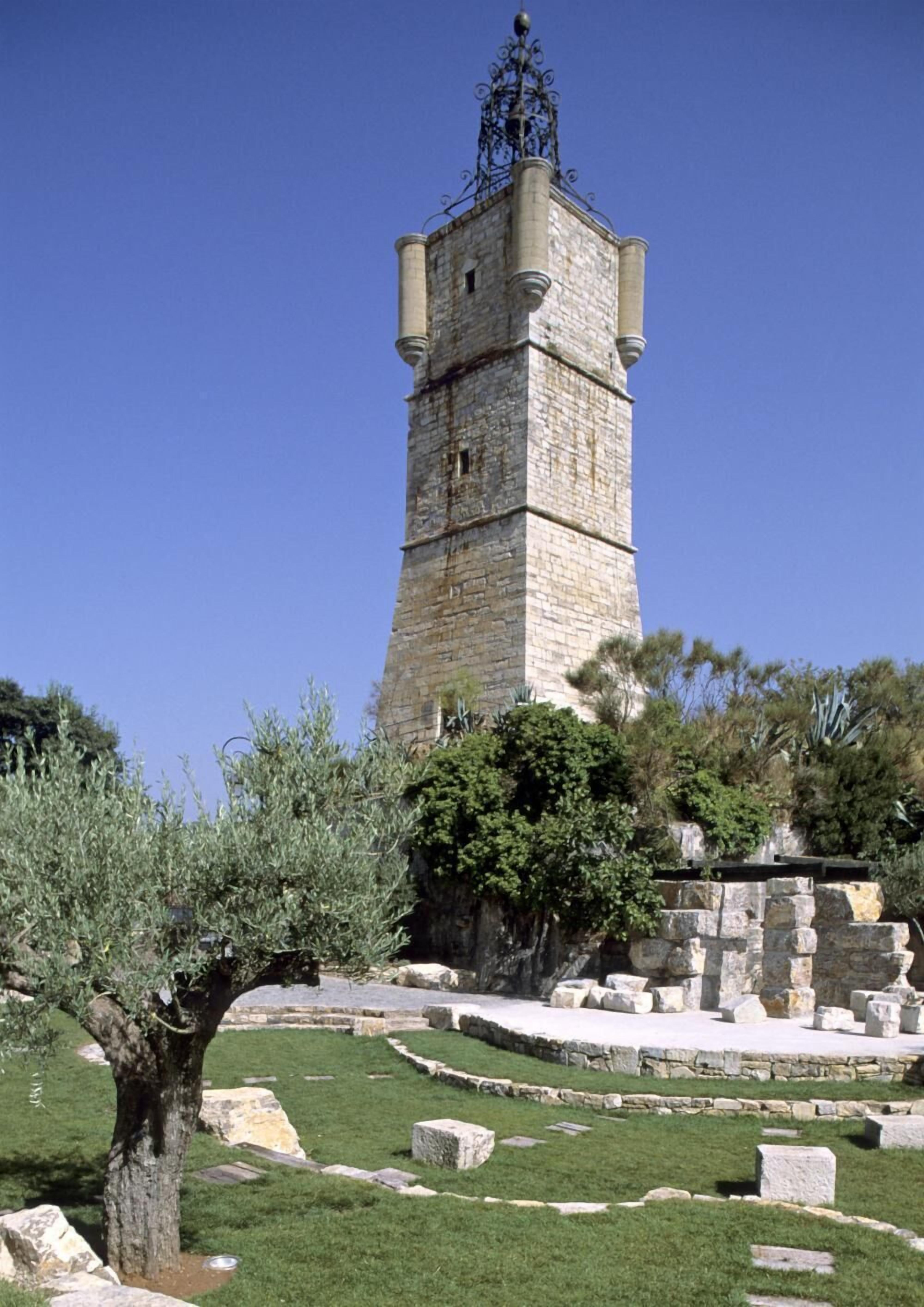 A stone tower with a metal spire, surrounded by a grassy area with scattered stones and a tree.