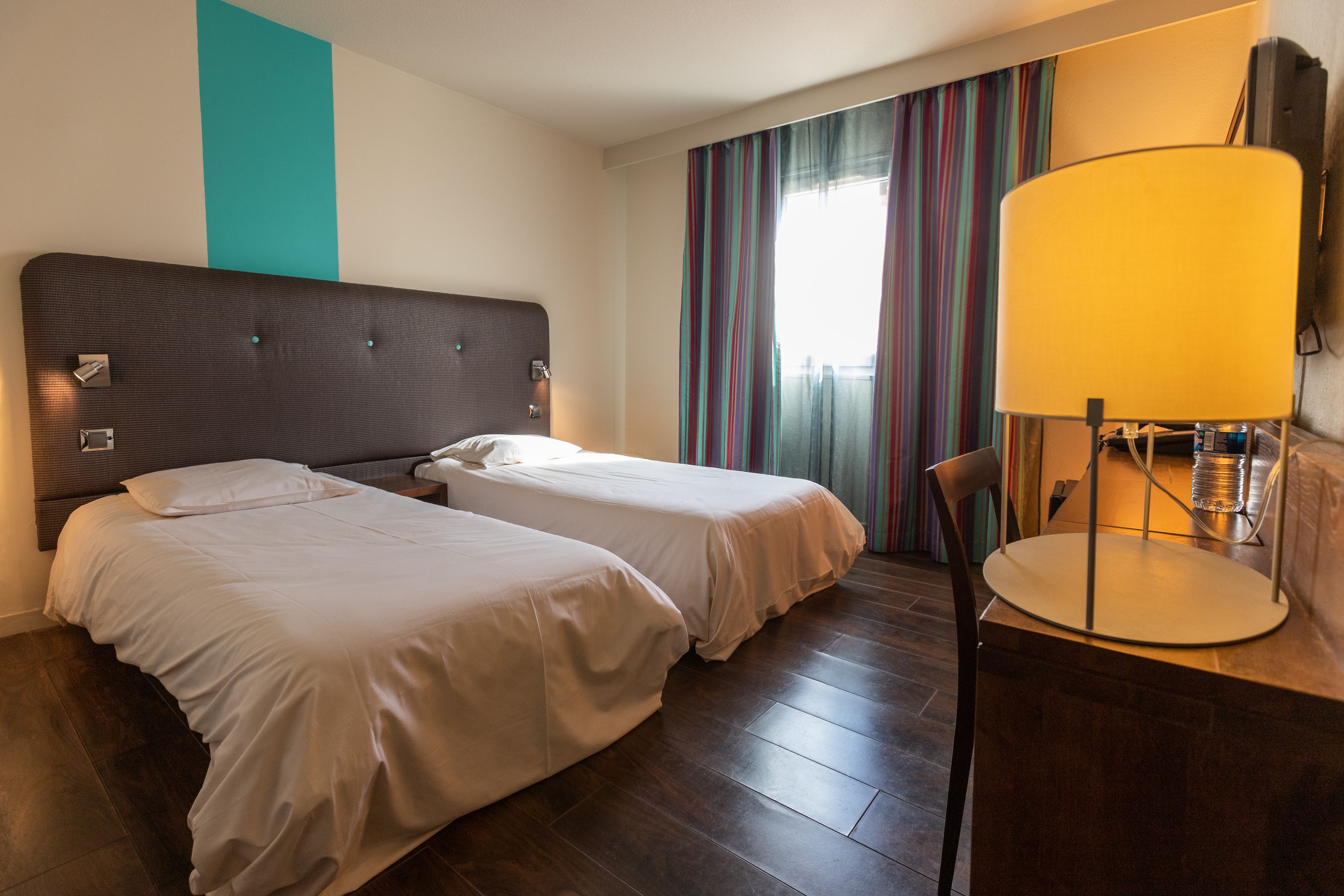 A hotel room with two beds, a wooden floor, a desk with a lamp, and a window with curtains.