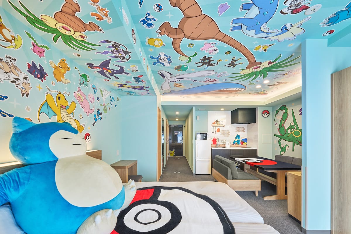 A room with a bed featuring a Pokemon-themed bedspread, a large plush toy, and a wall decorated with various Pokemon characters.