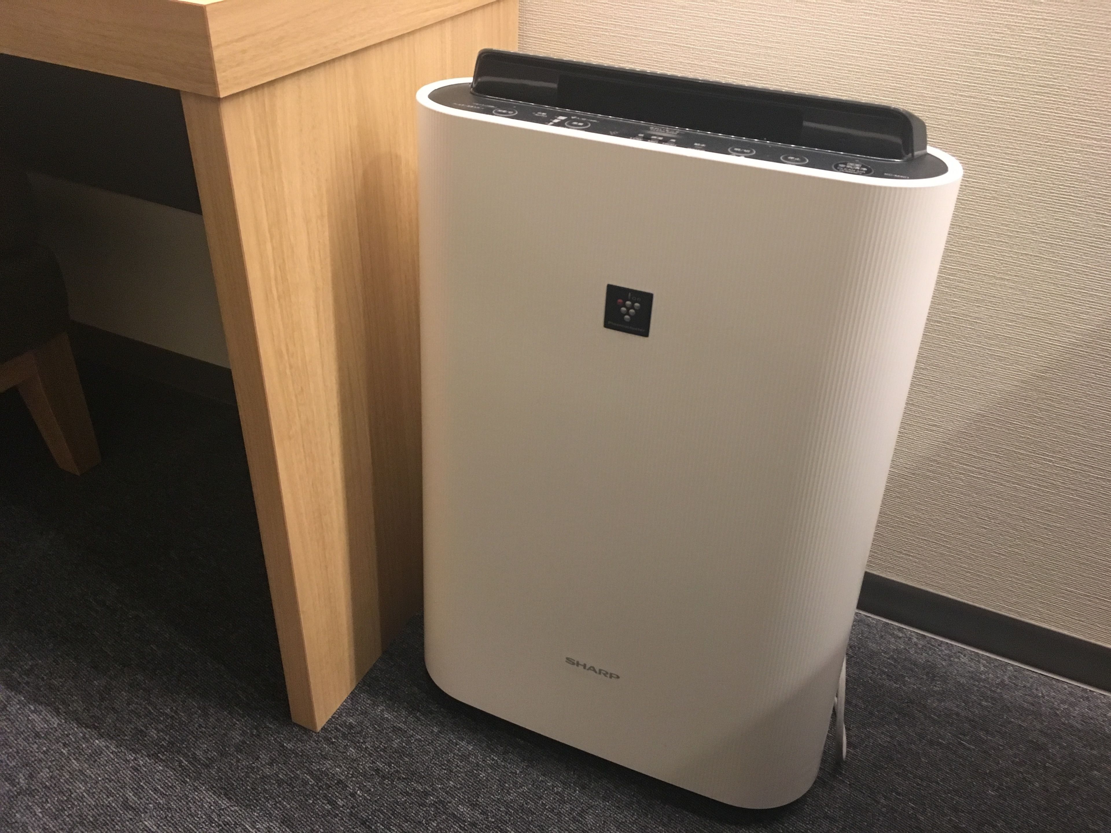A white, modern air purifier with a control panel and a small display, placed on a carpeted floor next to a wooden table.