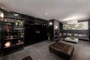 Una sala de estar moderna con un sofá de tonos oscuros, un reposapiés de cuero y una mesa de centro. Se observa una estantería con varios objetos decorativos y un mural grande en la pared.