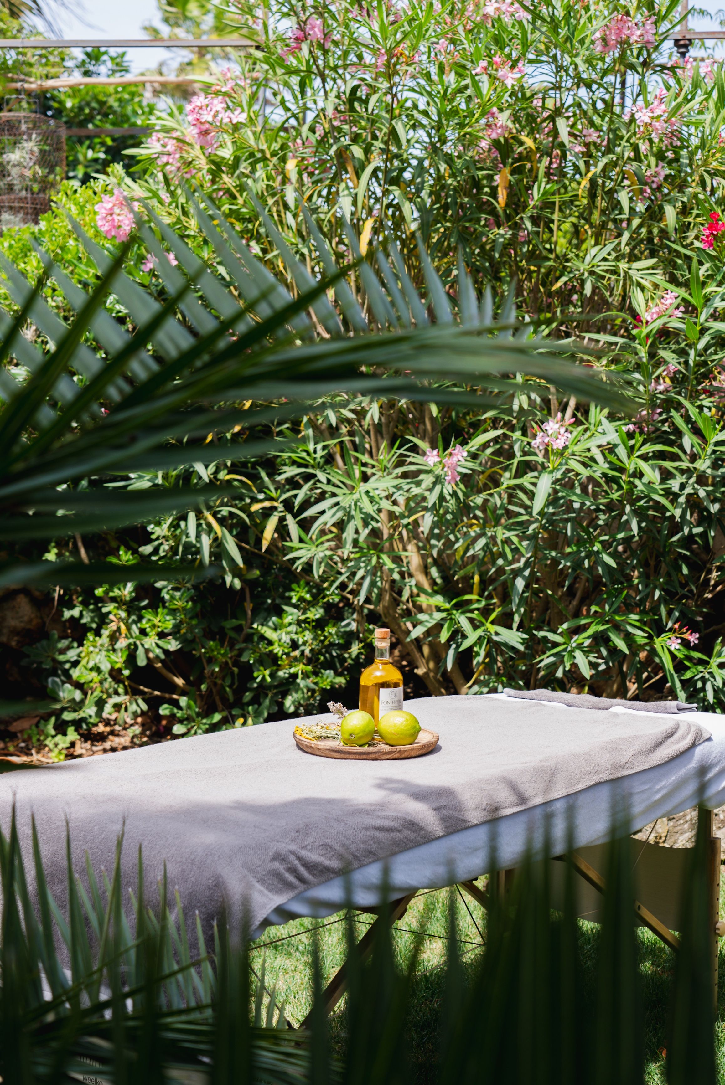 Mesa al aire libre con una botella y limones sobre una tabla de madera, rodeada de follaje y flores.