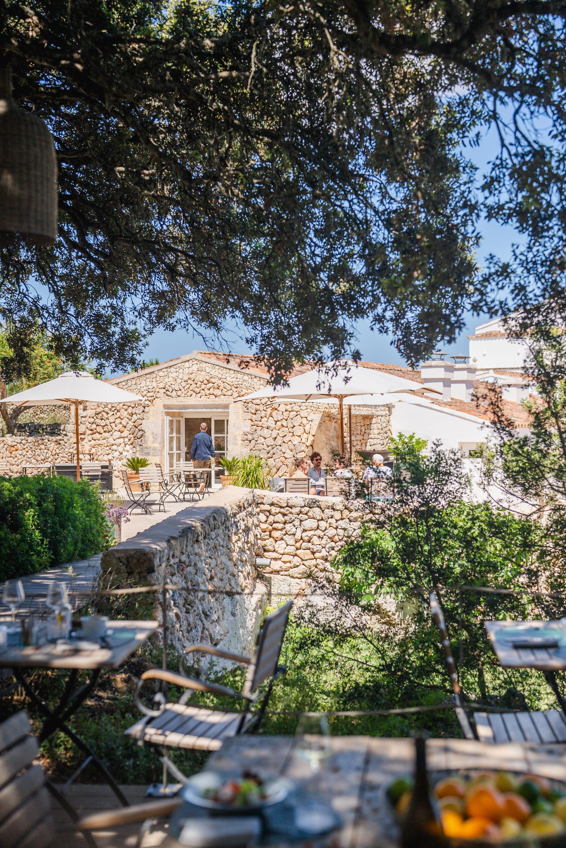 An outdoor seating area with stone walls and tables, surrounded by greenery and trees.