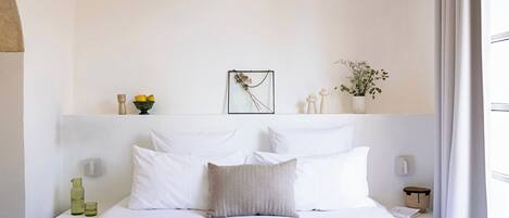 A minimalist hotel room with a bed, white walls, a framed picture, and a small plant.