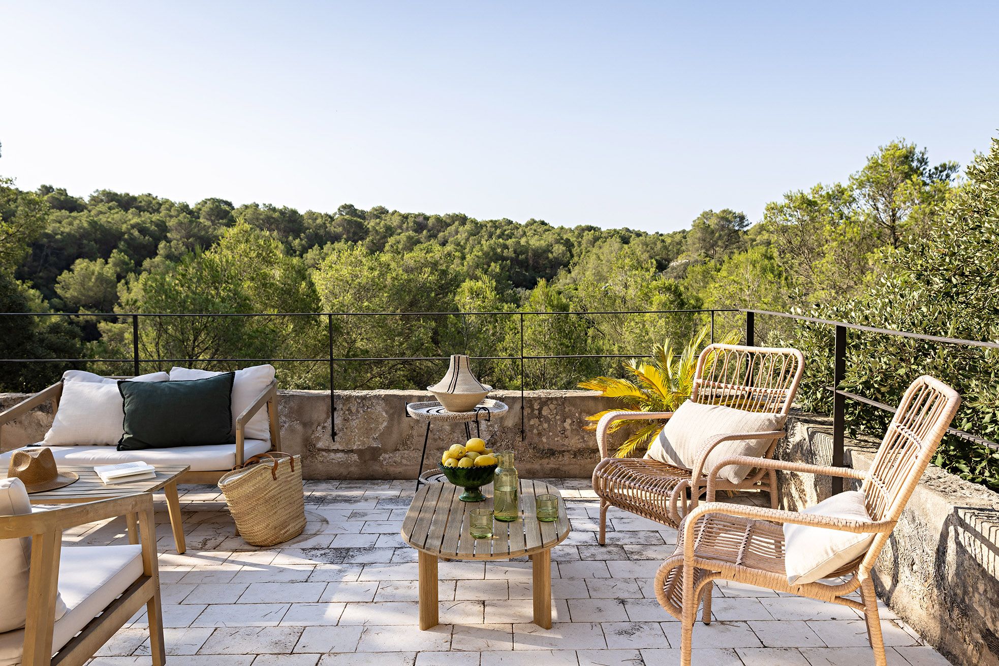 An outdoor seating area with wicker furniture, a round table with a bowl of lemons, and a view of a forested hillside.