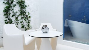 A white table with a glass top and a metal pitcher, surrounded by white chairs, in a room with a white wall and a blue bathtub visible through a glass door.
