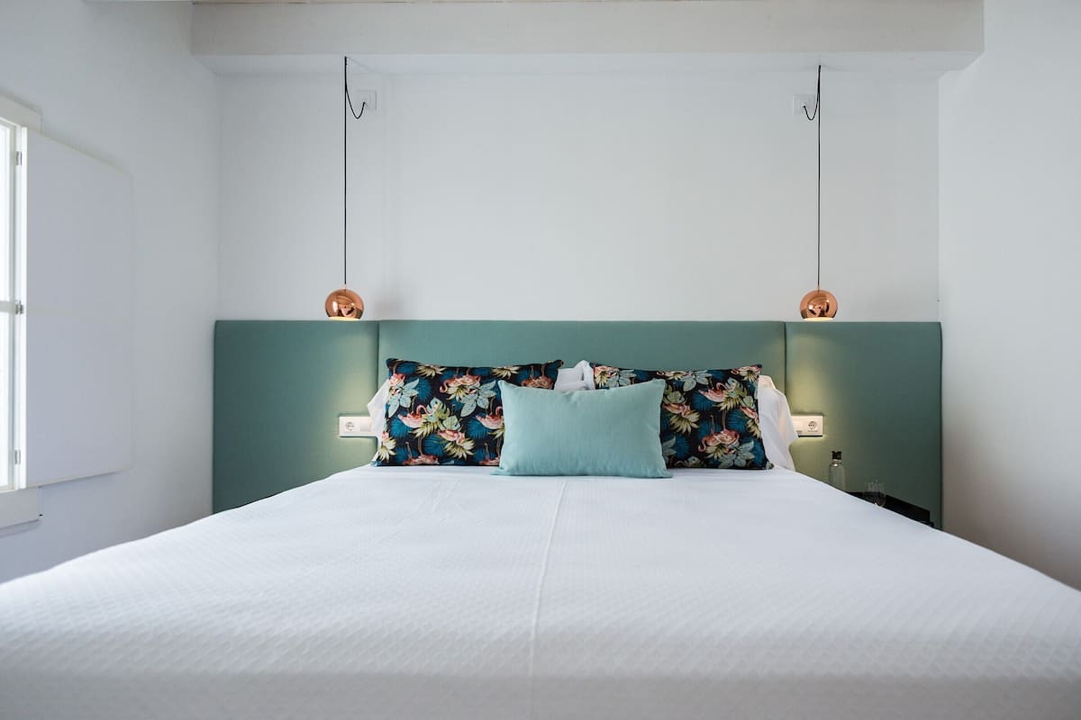 Un lit bien fait, avec une tête de lit à motifs floraux et deux lampes fixées au mur.
