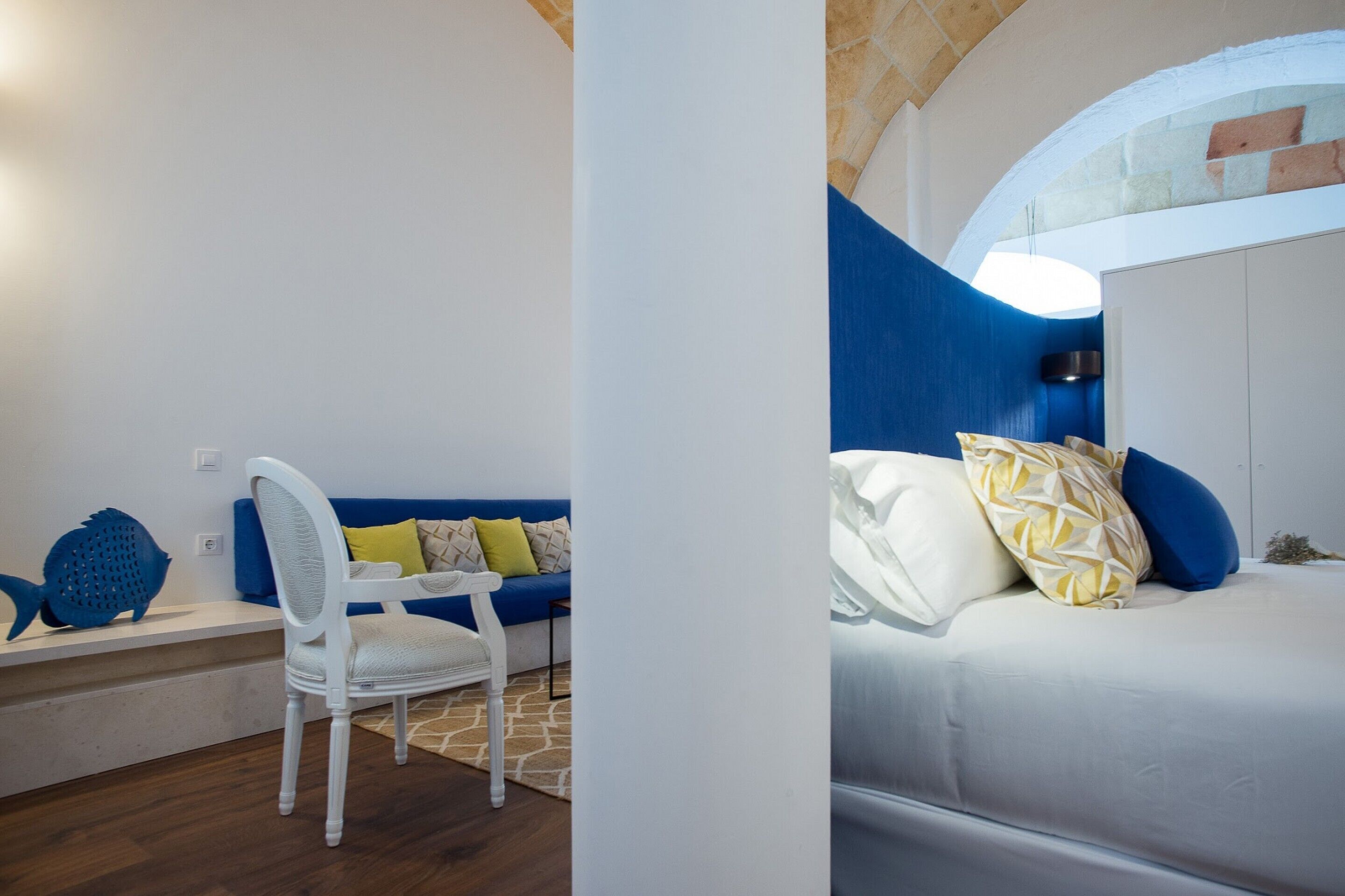 A room with a white bed, blue and yellow pillows, a white chair, and a blue sofa.