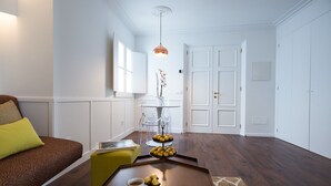 A modern living room with a brown sofa, a yellow ottoman, a coffee table with a cup and pastries, a clear glass table with flowers, and a pendant light.