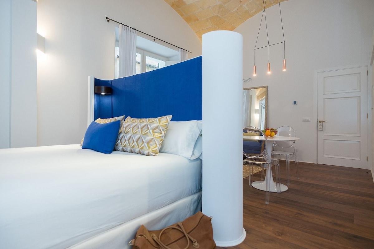 Une chambre moderne avec un grand lit, un mur d’accent bleu, une colonne blanche, une petite table avec des fruits et une chaise transparente.