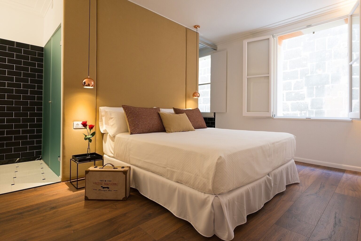 A hotel room with a bed, a nightstand, a suitcase, a vase with a rose, and a window with a view of a brick wall.