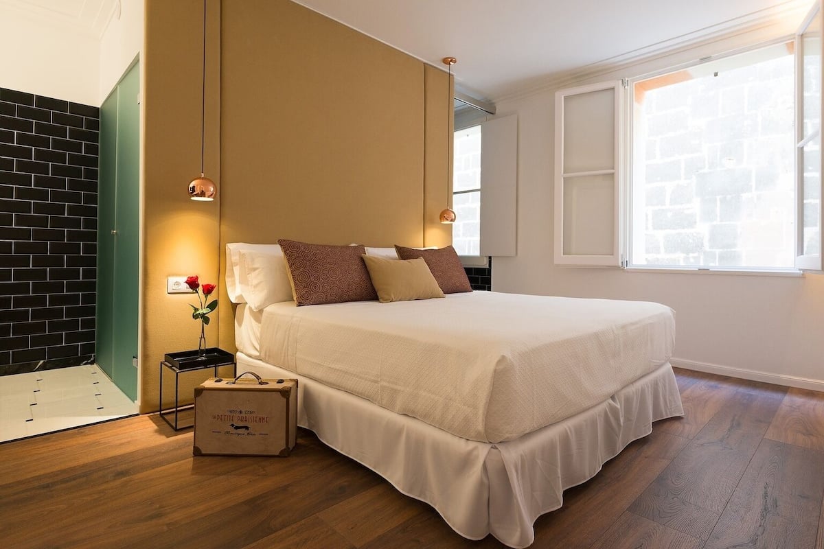 Une chambre d’hôtel avec un lit, une table de chevet, une valise, un vase contenant une rose et une fenêtre donnant sur un mur de briques.
