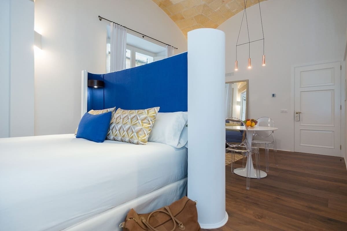 Une chambre moderne avec un grand lit, un mur d’accent bleu, une colonne blanche, une petite table avec des fruits et une chaise transparente.