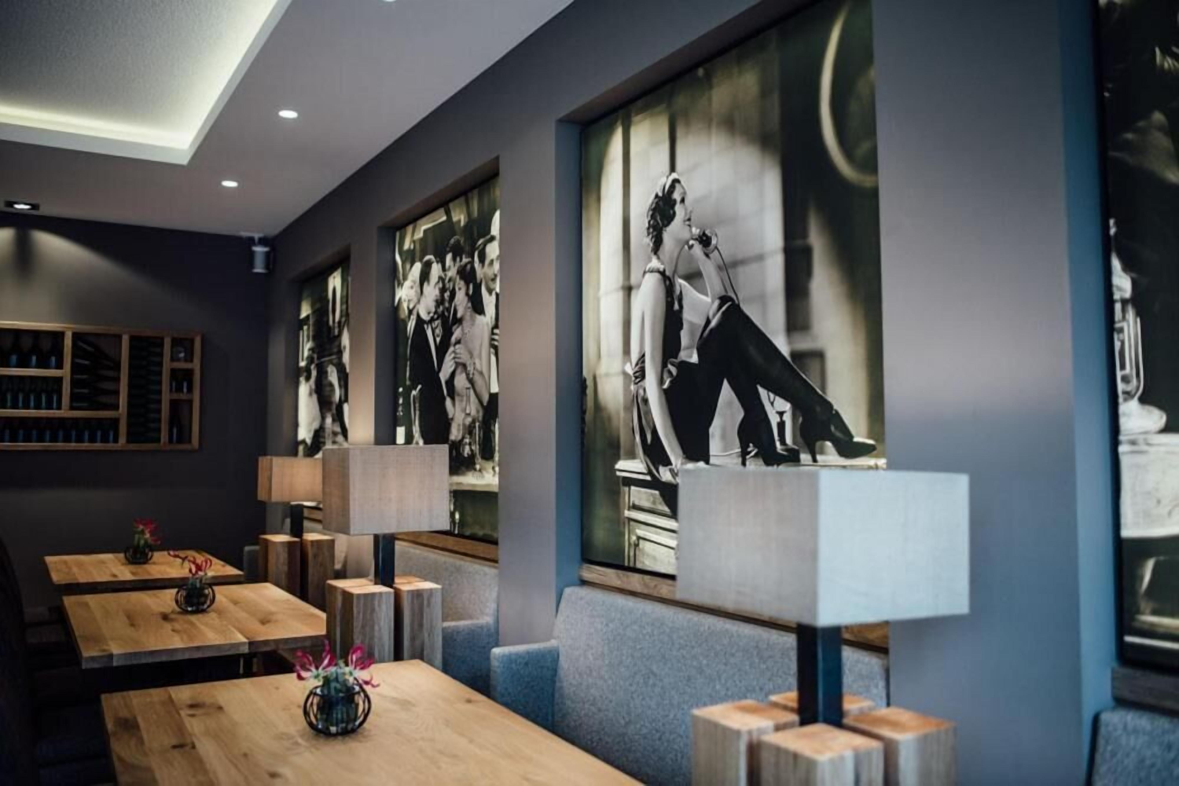 Een modern interieur met houten tafels en blauw gestoffeerde zitplaatsen, zwarwitfoto’s aan de muur en een plafond met inbouwverlichting.