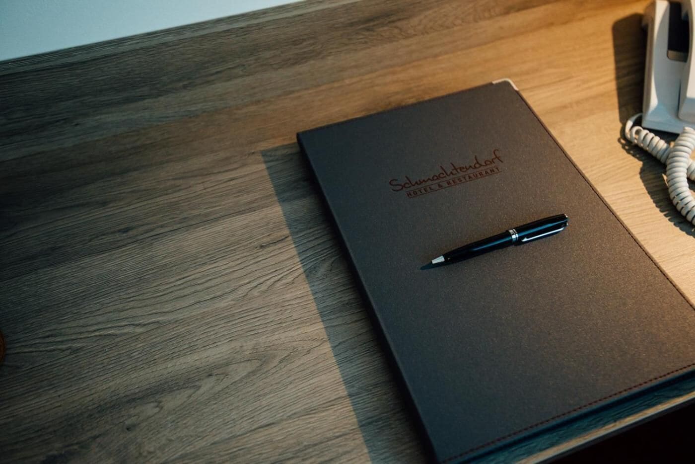 Un libro de tapa de cuero marrón oscuro titulado "Schmuckendorf Hotel & Restaurant" sobre una superficie de madera, con un bolígrafo y un teléfono.