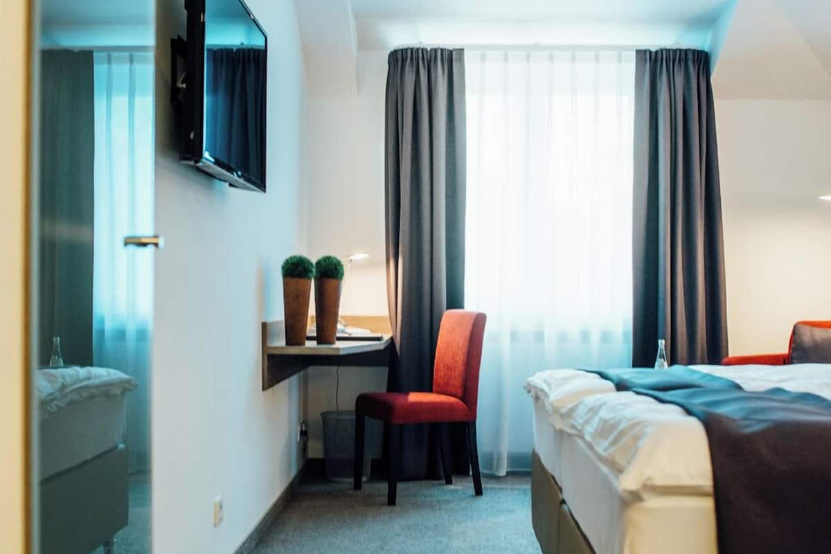 Habitación de hotel con una cama, un escritorio con silla, un televisor en la pared y una ventana con cortinas.