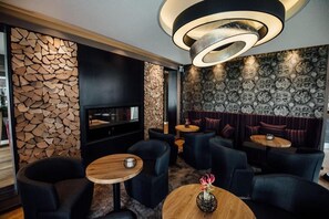 A modern lounge area with a stone wall, a fireplace, and a patterned wall. There are several seating areas with round tables and chairs. - Hotel und Restaurant Schmachtendorf (Oberhausen)
