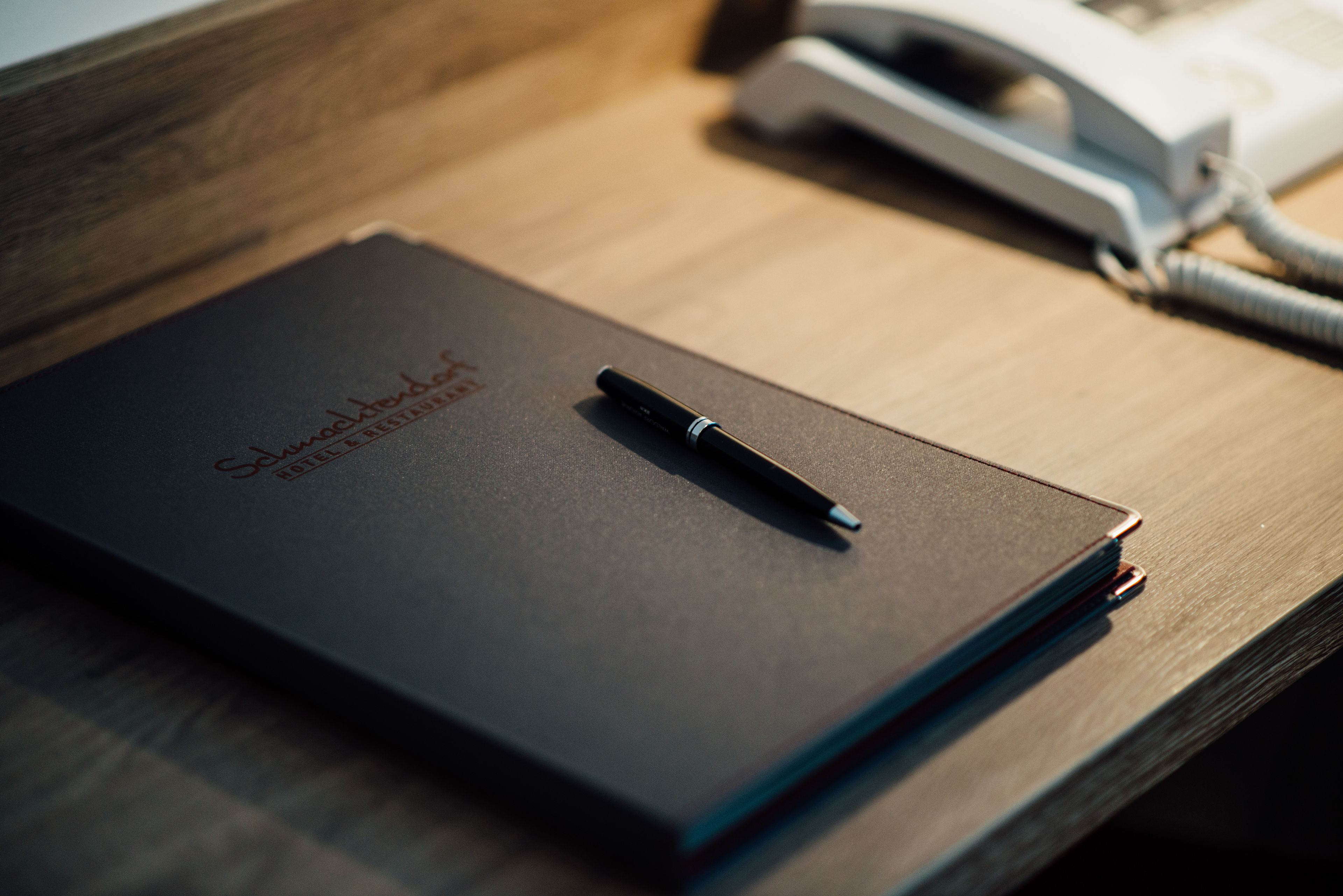 A dark-colored notebook with a pen on a wooden surface, accompanied by a white telephone.