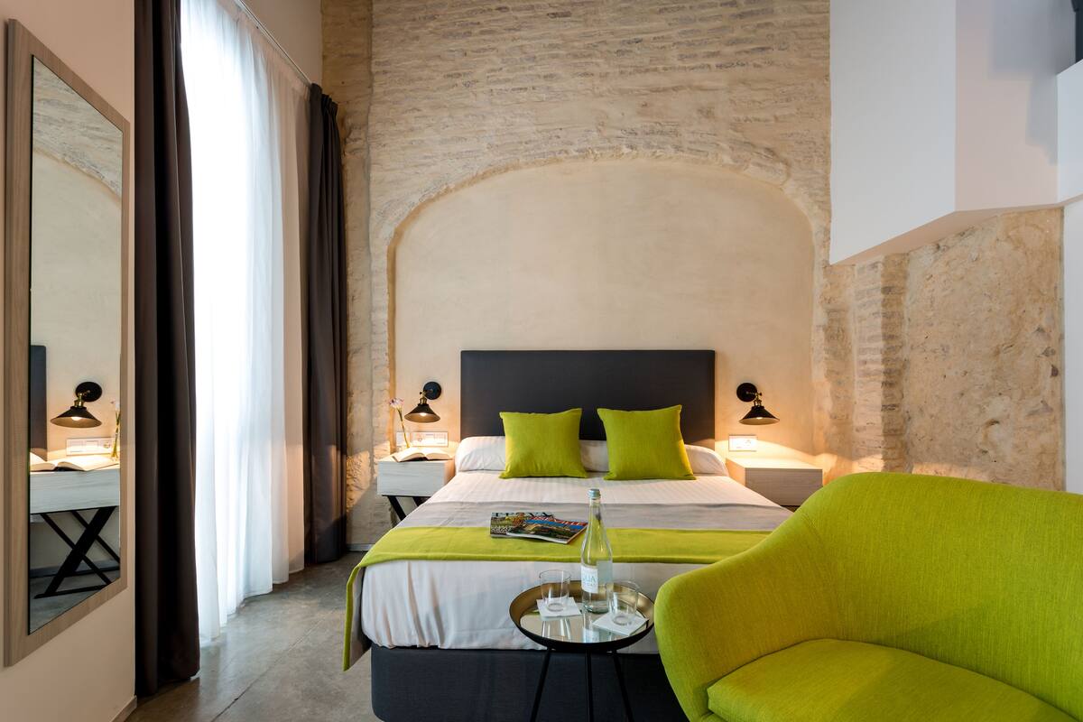 Een hotelkamer met een groot bed, groene kussens, een nachtkastje met een lamp en een felgroene fauteuil.