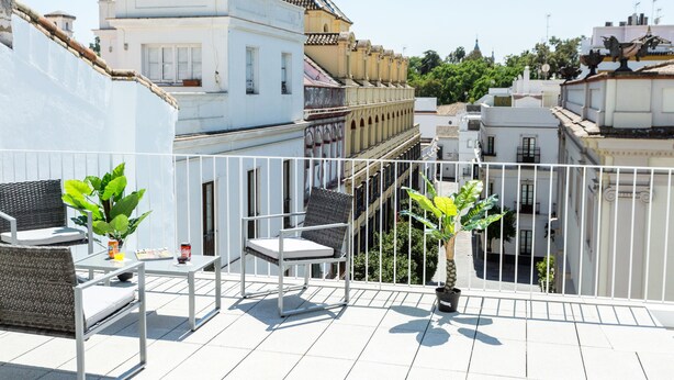 Un balcone con ringhiera bianca, un tavolino, una sedia e due piante in vaso.