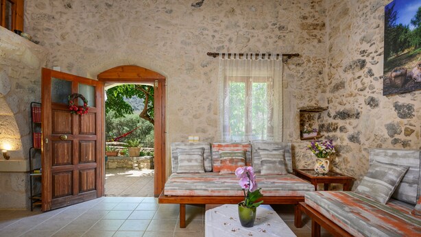 A stone-walled room with a wooden door, a sofa set, a small table with a vase, and a window with a view of greenery. - Pagonas Traditional House (Apokoronas)