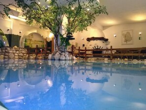 An indoor pool with a tree and rocks, wooden benches, and a stone wall. - Schlossberghof Marzoll (Bad Reichenhall)