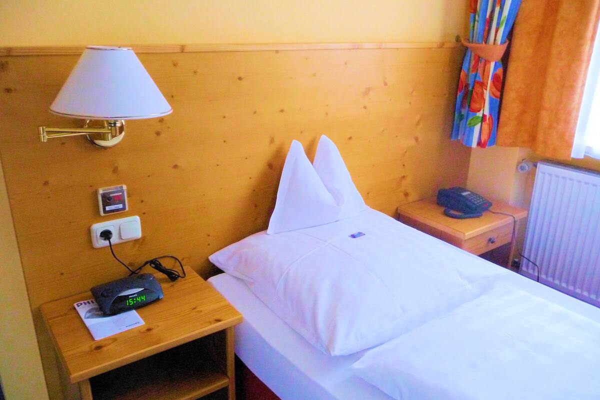 Habitación de hotel con cabecera de madera, lámpara de noche, mesita con teléfono, reloj digital y toma de corriente.
