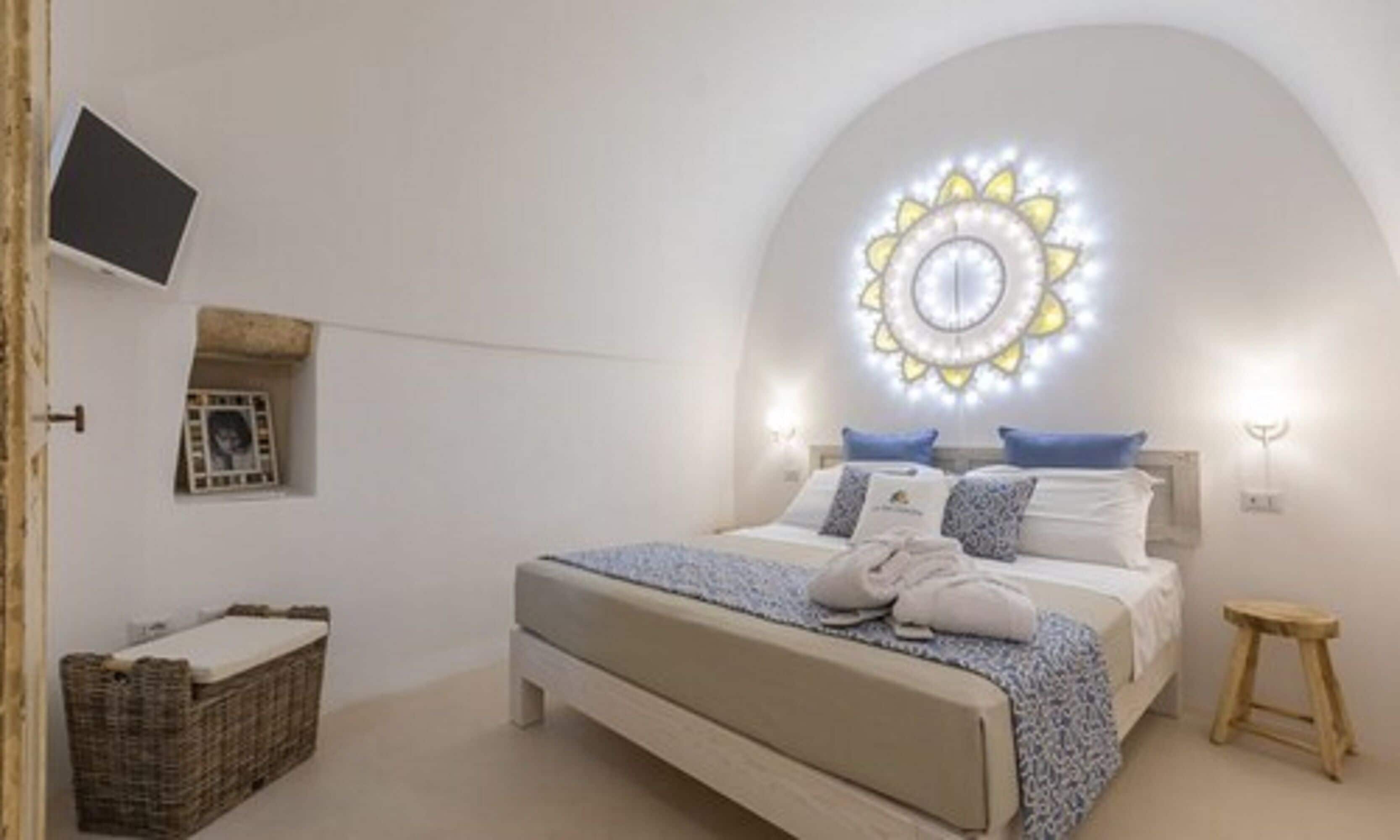 Un dormitorio con una cama, un televisor montado en la pared, una lámpara decorativa y un taburete pequeño.