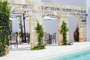 Un área junto a la piscina con arcos de piedra, mobiliario exterior y abundante vegetación.