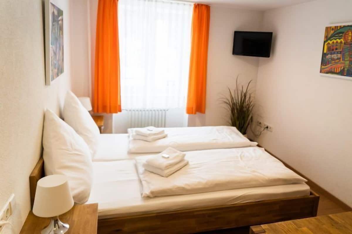 A hotel room with two beds, a TV mounted on the wall, and a window with orange curtains.