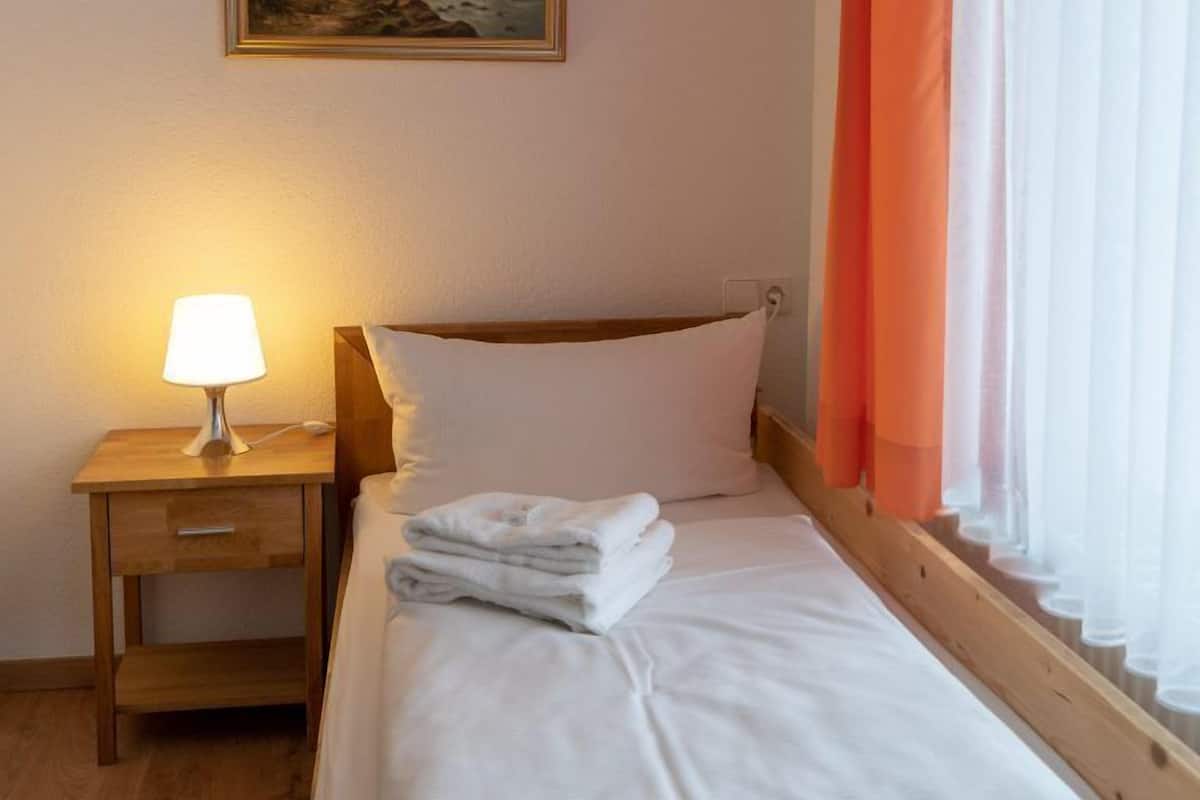 A neatly made bed with white linens and a folded towel, a bedside lamp, and a window with orange curtains.
