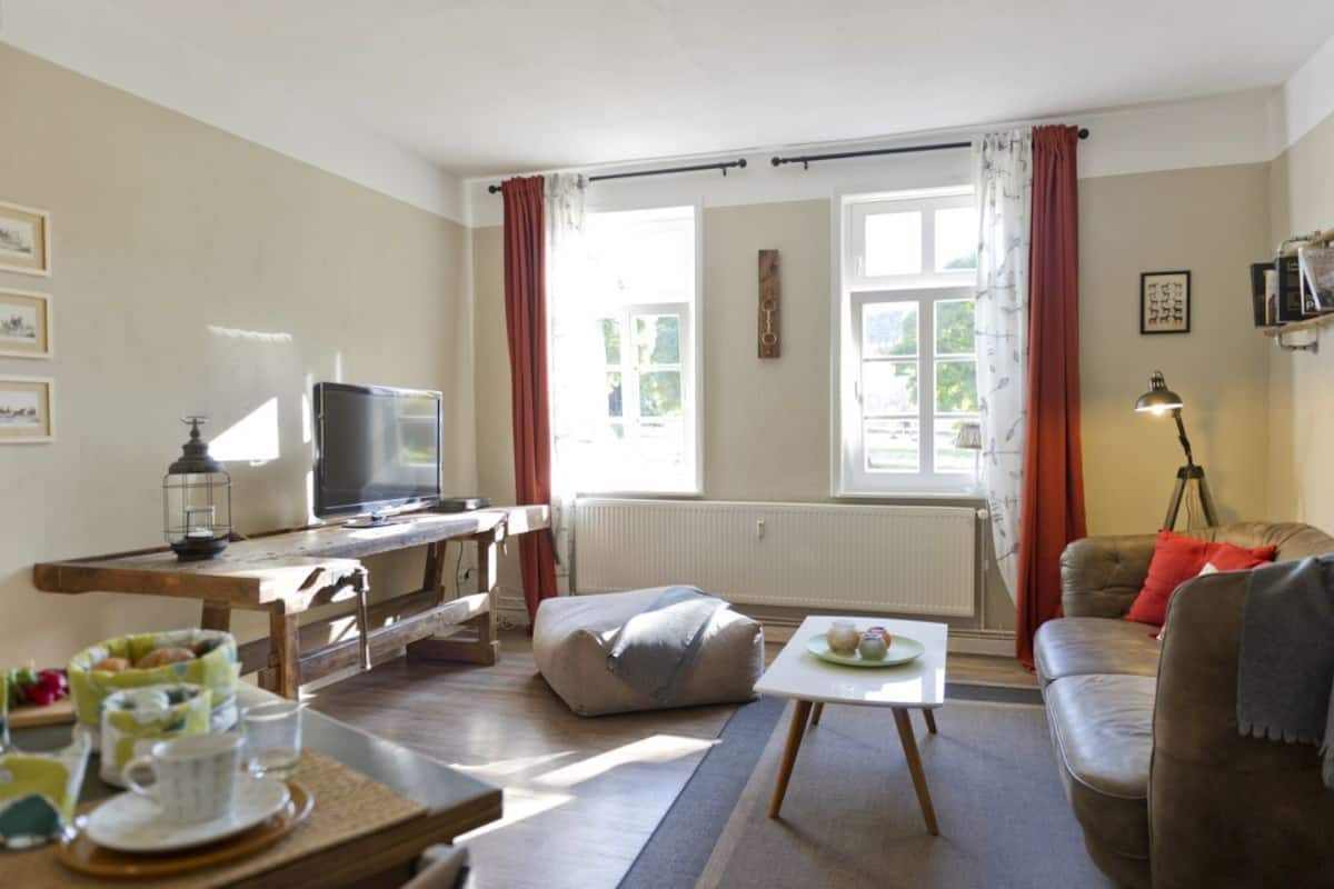 Uma sala de estar acolhedora, com televisão, mesa de jantar com abajur, sofá e janela com cortinas.