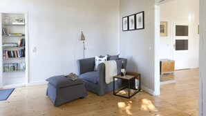 A modern living room with a grey sofa, a small table with a bottle and glass, and a bookshelf filled with books. - Gutshof Fürstenberg (Fürstenberg)