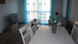 A modern living room with a white dining table, a TV on a wooden stand, and a vase with flowers. - By The Sea II (Limassol)
