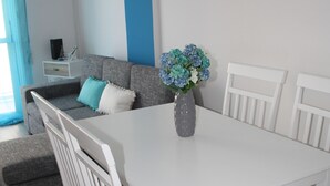 A dining area with a white table and chairs, a grey sofa with blue cushions, and a vase with flowers. - By The Sea II (Limassol)