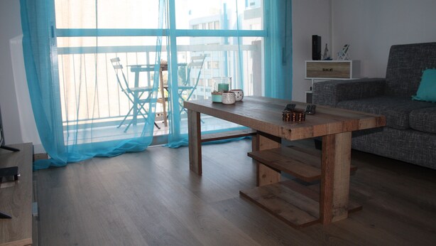 A room with a wooden table, a sofa, a balcony with chairs, and a view of buildings through sheer curtains. - By The Sea II (Limassol)