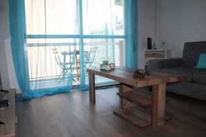 Une pièce avec une table en bois, un canapé, un balcon avec des chaises et une vue sur les immeubles à travers des rideaux légers.