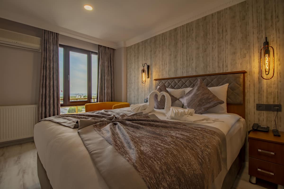 A hotel room with a large bed, a bedside table, a chair, a window with curtains, and a wall-mounted light fixture.
