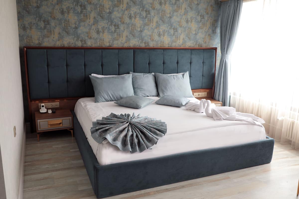 A large bed with a dark headboard, multiple pillows, and a decorative pillow on the bed. There is a wooden nightstand with a phone and a small drawer. The room has a light-colored wooden floor and a textured wall.