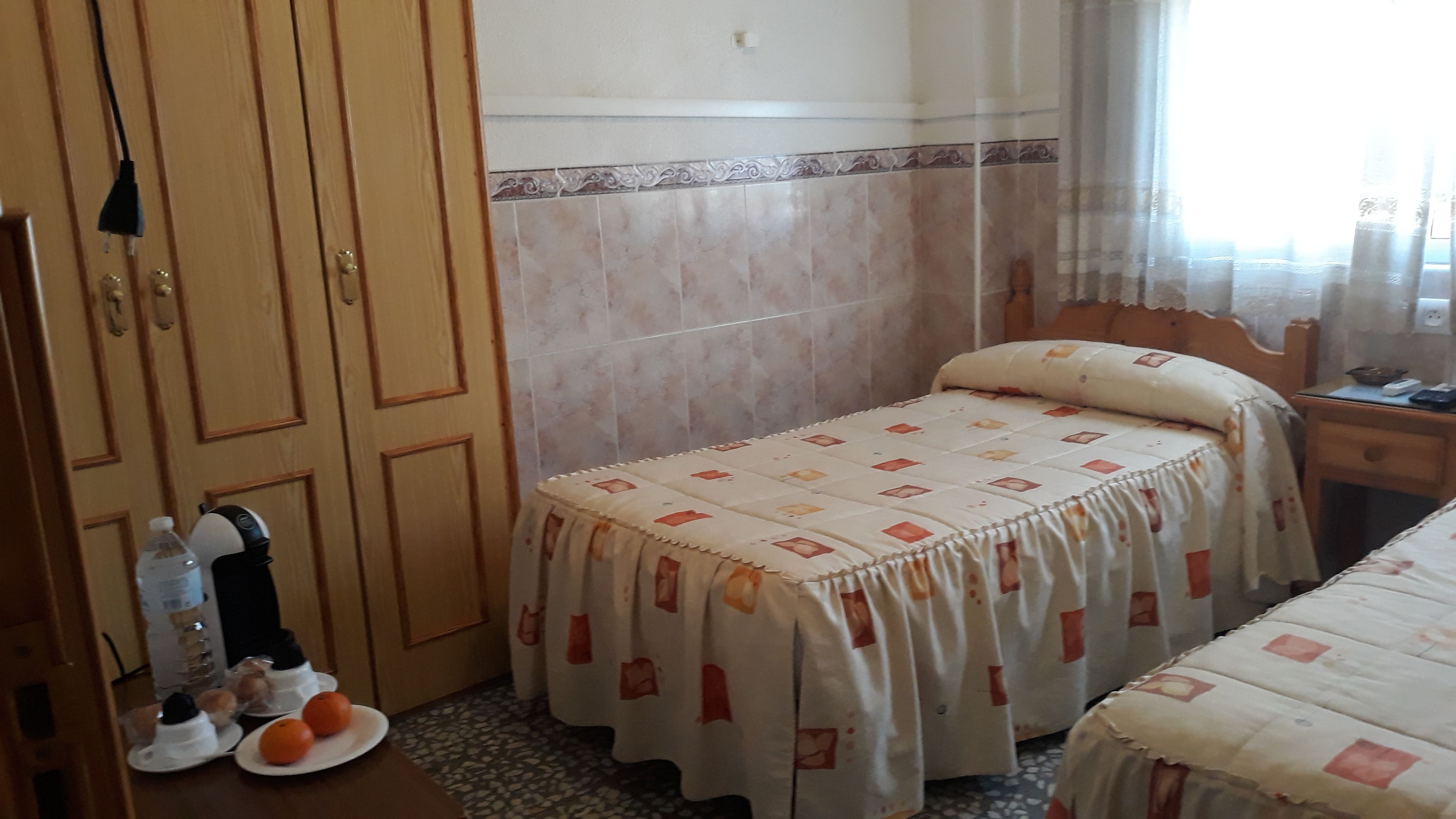 A bedroom with a bed, a nightstand, a wooden wardrobe, and a small table with a water bottle and oranges.