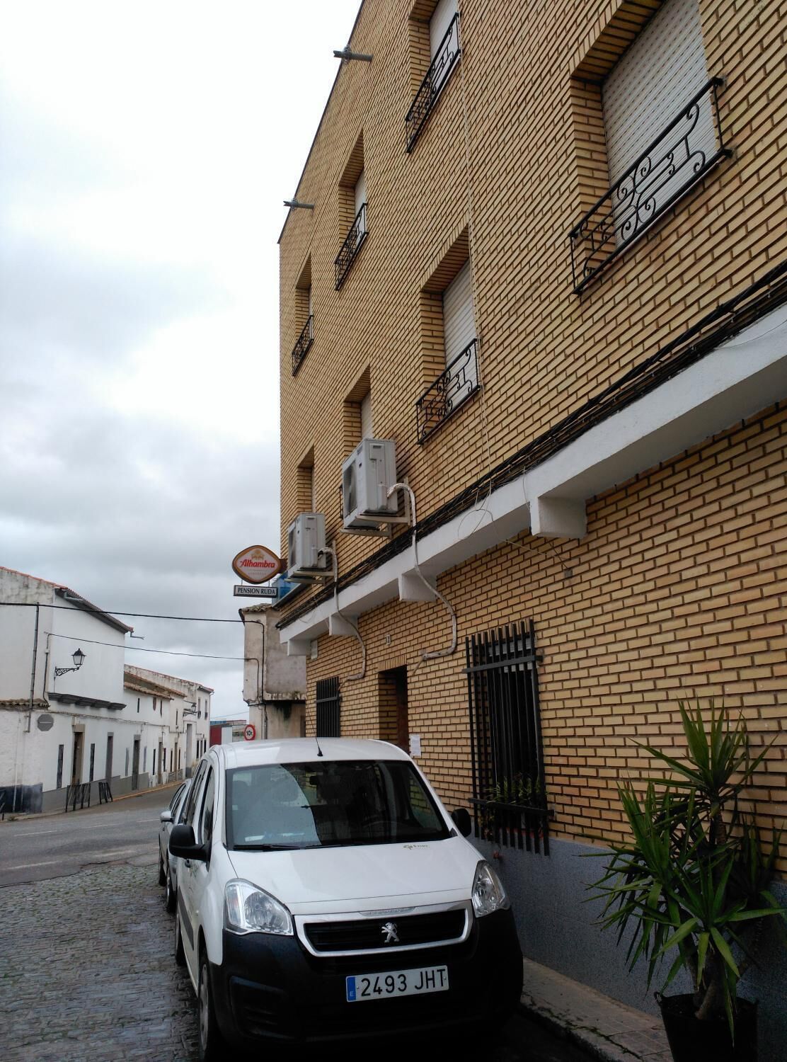 A white Peugeot car parked on a cobblestone street in front of a brick building with air conditioning units and a sign that reads "Alhambra Pension Real".