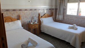 A hotel room with two single beds, a small bedside table with toiletries, a window with curtains, and a tiled wall with a decorative border. - Pension Ruda (Hinojosa del Duque)