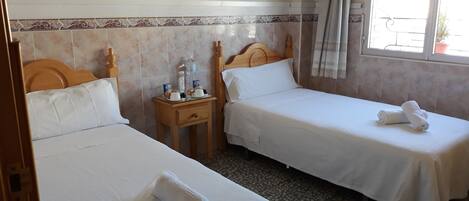 A hotel room with two single beds, a small bedside table with toiletries, a window with curtains, and a tiled wall with a decorative border.