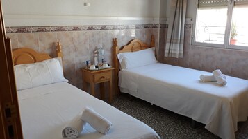 Habitación de hotel con dos camas individuales, una mesita de noche pequeña con artículos de aseo, una ventana con cortinas y una pared de azulejos con un borde decorativo.